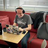 Constantin with headphones sitting at a table in an office break area, smiling while looking at a laptop covered in various tech and programming stickers. Large windows show a cityscape view of Munich office buildings in the background.