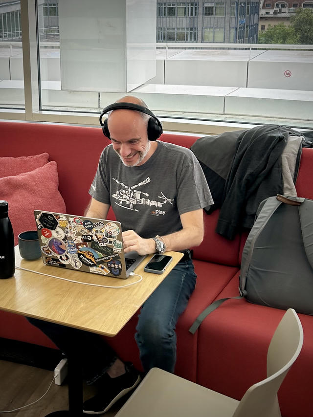 Constantin with headphones sitting at a table in an office break area, smiling while looking at a laptop covered in various tech and programming stickers. Large windows show a cityscape view of Munich office buildings in the background.