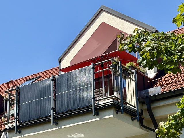 A multi-story residential building with red clay tile roofing under a clear blue sky. The upper level features a balcony with metal railings and solar panels mounted as balcony power plant system (Balkonkraftwerk). Above the balcony is a triangular dormer window with cream and reddish-brown colored walls, topped by a red awning. Green leafy vines or foliage are visible growing around the right side of the building.
