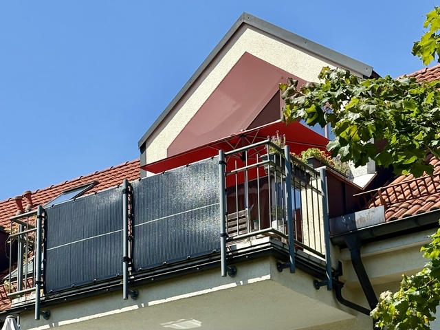 A multi-story residential building with red clay tile roofing under a clear blue sky. The upper level features a balcony with metal railings and solar panels mounted as balcony power plant system (Balkonkraftwerk). Above the balcony is a triangular dormer window with cream and reddish-brown colored walls, topped by a red awning. Green leafy vines or foliage are visible growing around the right side of the building.