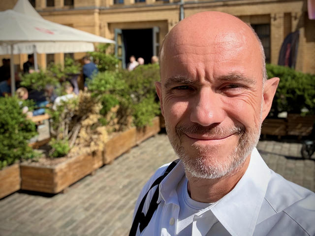 Constantin, wearing a white shirt, smiles at the camera during an outdoor event. Behind him is a courtyard with wooden planters containing green plants, a white tent, historic stone buildings, and other event attendees enjoying the sunny day.