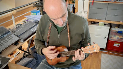 Constantin, wearing an olive green zip-up, sits at his desk playing a small wooden ukulele. He is focused on the instrument as his hands position the strings. Behind him is a wooden desk with a keyboard, music equipment, and storage cabinets.