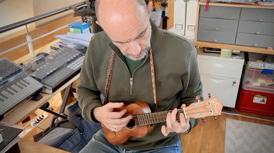 Constantin, wearing an olive green zip-up, sits at his desk playing a small wooden ukulele. He is focused on the instrument as his hands position the strings. Behind him is a wooden desk with a keyboard, music equipment, and storage cabinets.