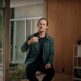 YouTube video screenshot showing a man in a dark green button-up shirt over a black t-shirt who sits casually in a modern living room with large windows showing a garden view. He appears to be speaking or presenting, with built-in shelving visible in the background and natural lighting from the windows.