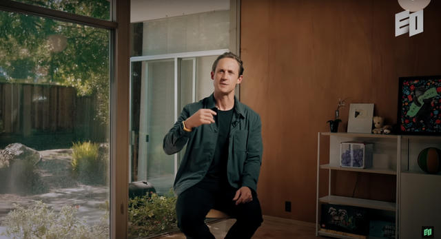 YouTube video screenshot showing a man in a dark green button-up shirt over a black t-shirt who sits casually in a modern living room with large windows showing a garden view. He appears to be speaking or presenting, with built-in shelving visible in the background and natural lighting from the windows.