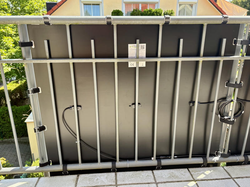 A solar panel mounted vertically on straps in a metal frame structure on a balcony. The panel has cables connecting to an adjacent panel and there are small identification labels visible. In the background, there is a yellow house with red roof tiles, green trees, and landscaping visible below the balcony.
