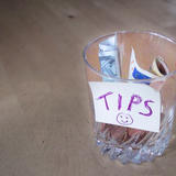 A glass jar labeled “TIPS” with a smiley face, filled with folded banknotes and coins, sitting on a wooden floor.