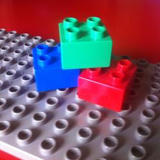 Three interlocking building blocks—blue, green, and red—stacked together on a grey studded baseplate against a red background.