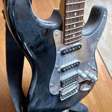 A black electric guitar with a contoured body rests against a wooden floor and wall. The guitar features a rosewood fretboard with metal frets, three black single-coil pickups arranged vertically on the pickguard, and a chrome bridge assembly at the bottom. A black fabric guitar strap is visible on the left side. The instrument is heavily covered in dust, indicating it has not been played in over a decade. The background reveals wooden flooring and a beige-colored rug.