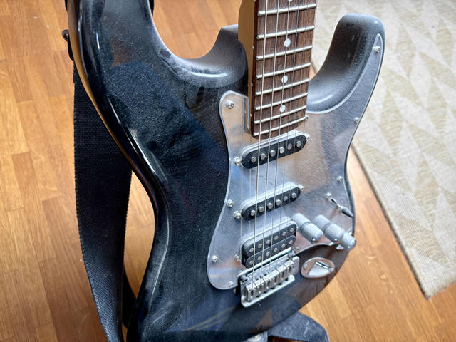 A black electric guitar with a contoured body rests against a wooden floor and wall. The guitar features a rosewood fretboard with metal frets, three black single-coil pickups arranged vertically on the pickguard, and a chrome bridge assembly at the bottom. A black fabric guitar strap is visible on the left side. The instrument is heavily covered in dust, indicating it has not been played in over a decade. The background reveals wooden flooring and a beige-colored rug.