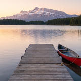 A calm mountain lake at dawn with a wooden jetty leading into the water and a red canoe moored beside it. Snow-dusted peaks and dark pine forests line the far shore under a soft pink and orange sky. No text or numbers appear anywhere in the image.